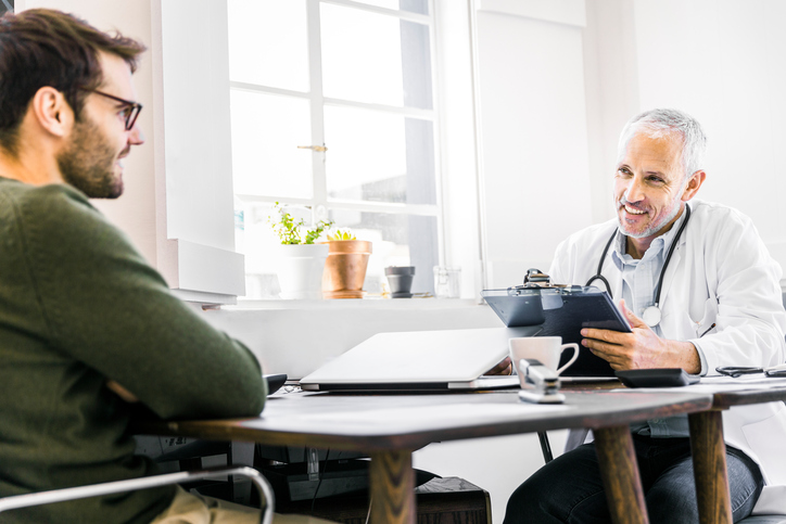 A photo of happy male doctor discussing with patient in clinic. Smiling medical professional is sitting at desk. Man is consulting healthcare practitioner in office.